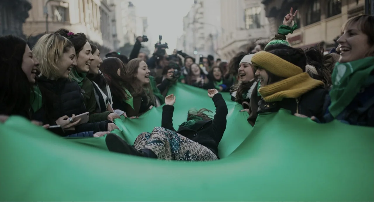 Belén (2025) movie still: medium — A group of women in a city street hold a large green banner aloft. A person lies back in the middle of the fabric…