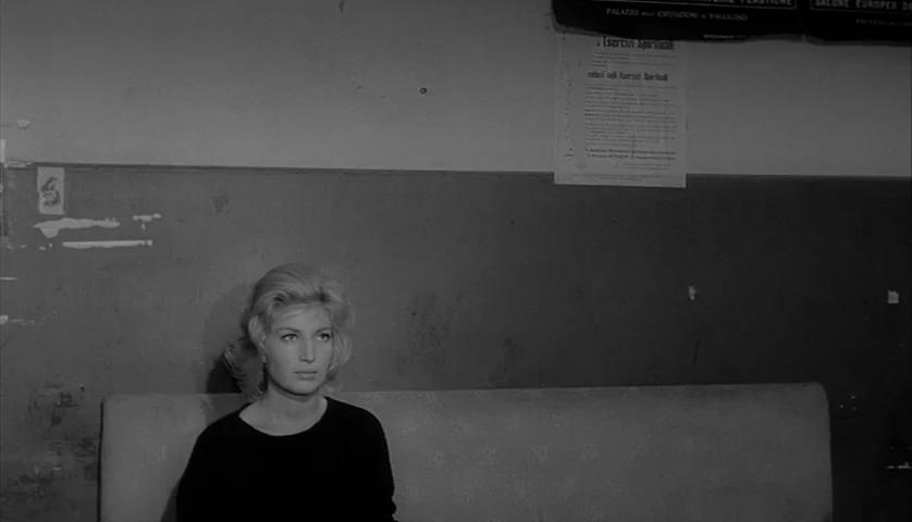 L’Avventura (1960) movie still: medium — A spare waiting area features a plain, worn wall with printed notices posted overhead. A blonde woman sits…