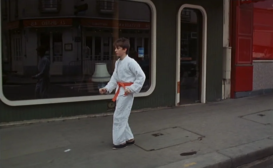 Kung Fu Master! (1988) movie still: fullbody — A sidewalk runs alongside a building front with a large display window. A teenage boy in a white…