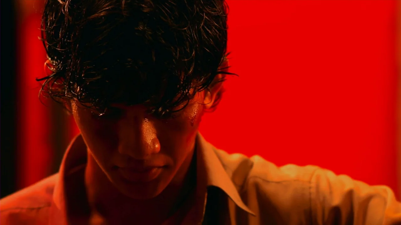 Merantau (2009) movie still: closeup — A close-up view of a young man with wet hair against a vibrant red backdrop. The man gazes downward while a…