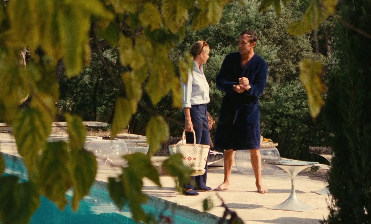 The Swimming Pool (1969) movie still: medium — A stone pool deck sits next to an in-ground swimming pool surrounded by greenery. A woman in trousers…