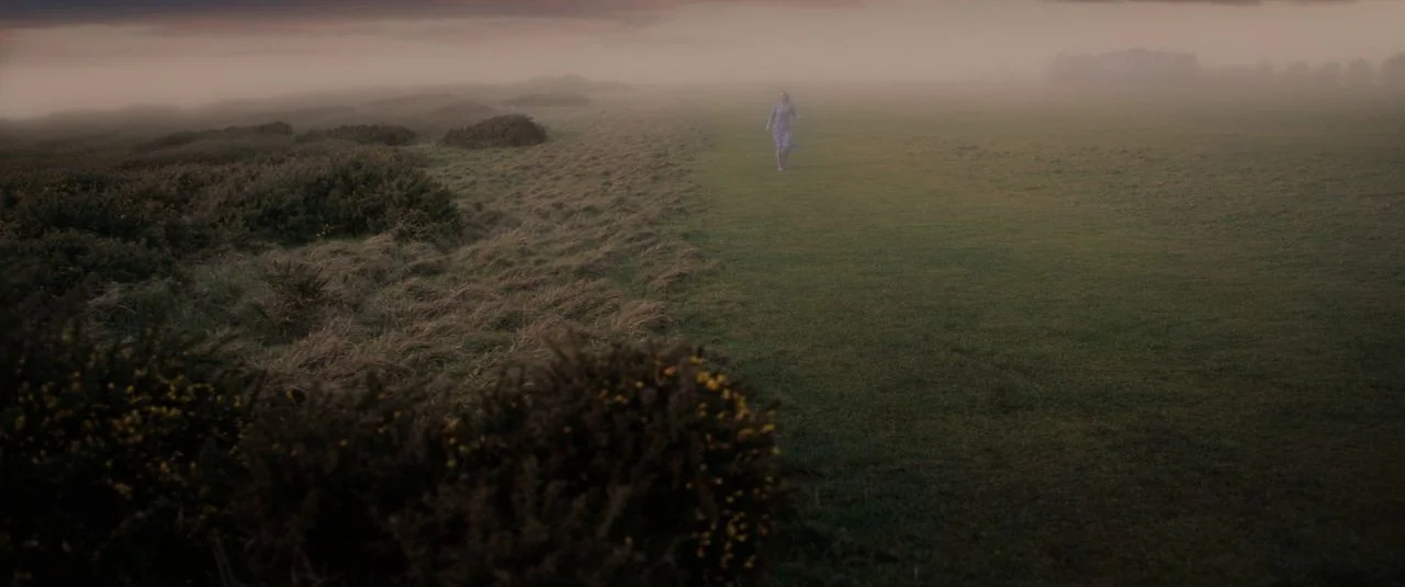 Rebecca movie still: wide — A vast, open landscape is covered in a thick, low-lying blanket of mist. A woman in a floral dress walks slowly along a…
