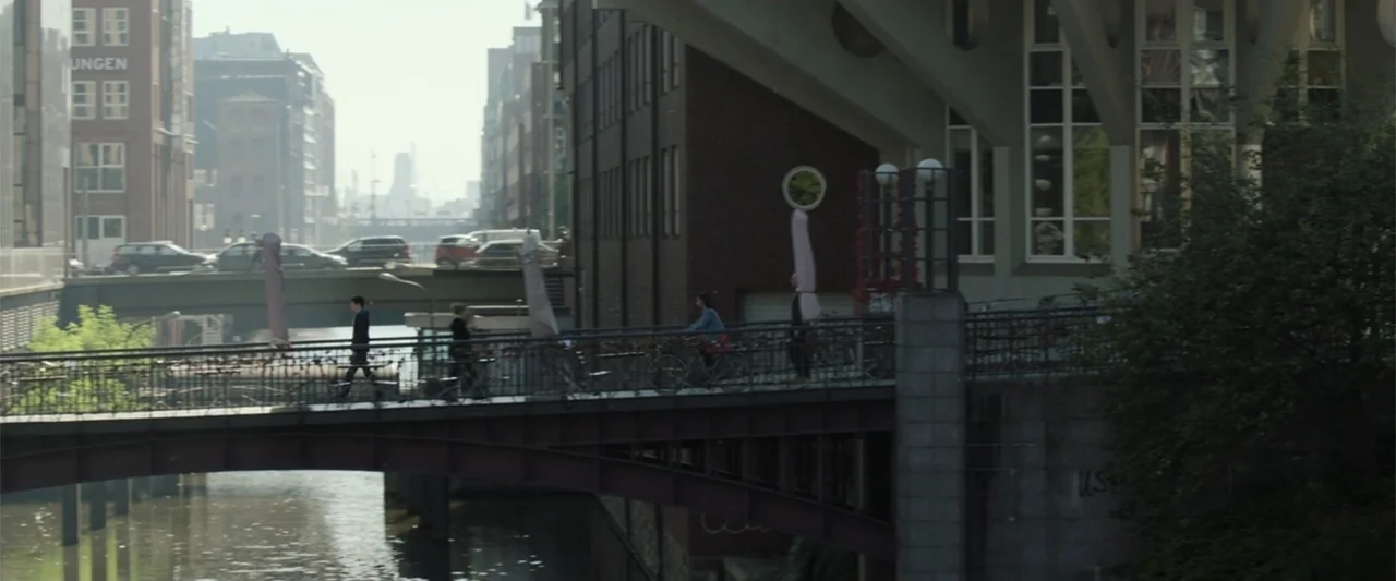 Charlie's Angels (2019) movie still: wide — A metal footbridge spans a narrow urban canal lined with brick buildings and cars. Pedestrians walk and…