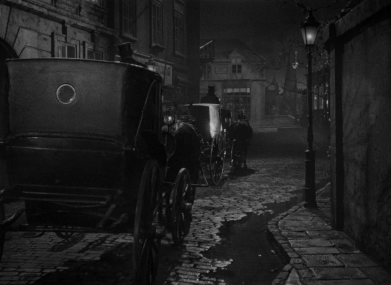 Letter From An Unknown Woman (1948) movie still: wide — A dark, narrow European-style cobblestone street features two horse-drawn carriages moving…