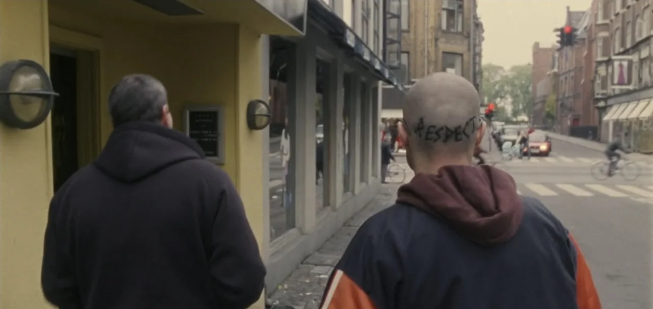 Pusher (1996) movie still: medium — An urban sidewalk runs alongside a building with large glass windows and street-level shops. Two men walk away…