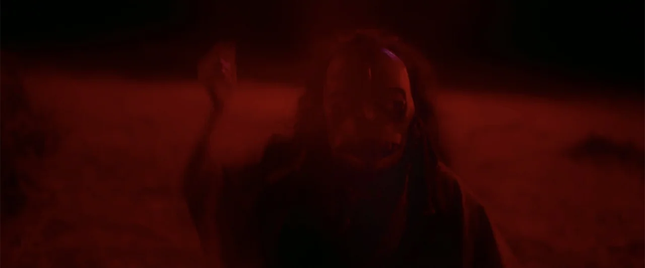 Evil Eye (2020) movie still: medium — An arid, dark exterior environment is illuminated by a piercing crimson light. A figure wearing a grotesque,…