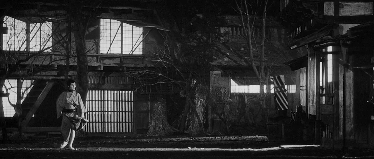 Yojimbo (1961) movie still: wide — A rural settlement featuring weathered wood buildings and paper-paneled sliding doors at night. A lone man moves…