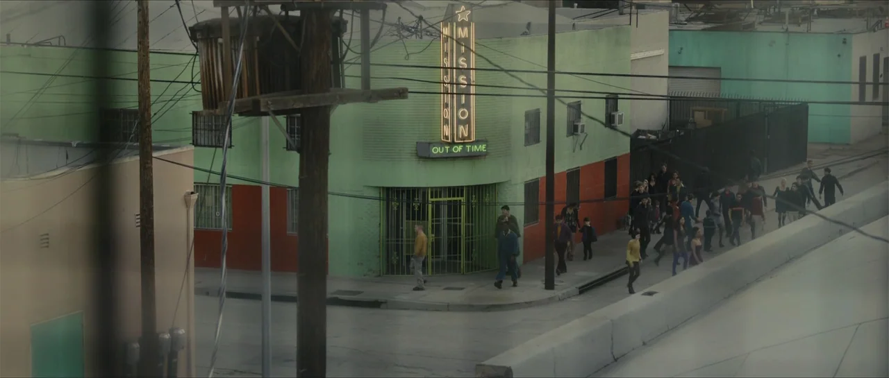 In Time (2011) movie still: wide — A corner building with a green exterior and a lit neon sign sits roadside. A diverse group of people walk along…