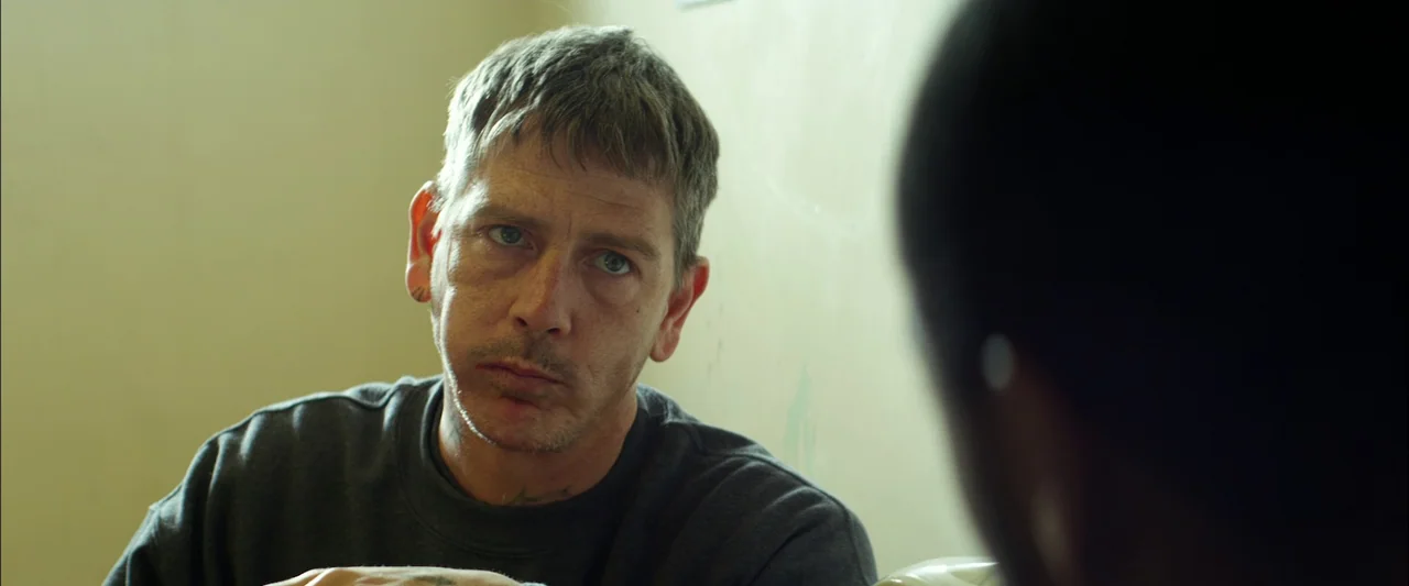 Starred Up (2013) movie still: medium — A man sits in a plain room with light beige walls. The man listens to another person whose back is to the…