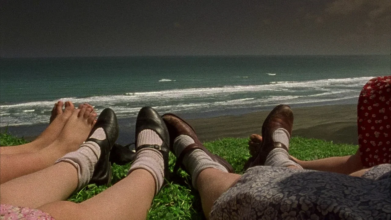 An Angel At My Table (1990) movie still: wide — A grassy hill overlooking the ocean during the day under a dark sky. Several pairs of legs and feet…