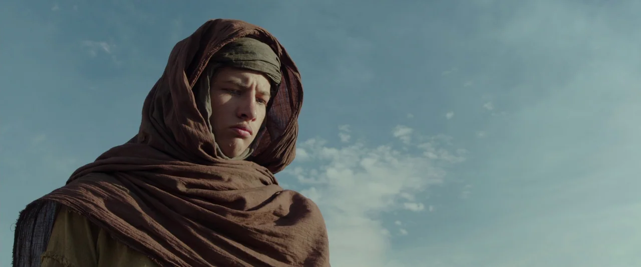 Last Days in the Desert (2015) movie still: medium — A man stands outdoors under a wide-open sky with light cloud cover. He wears traditional brown…