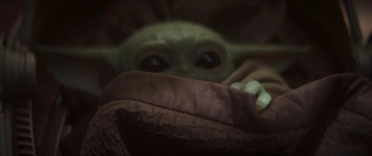 The Mandalorian (2019) movie still: closeup — A small green humanoid creature sits inside a rounded metallic transport pod. The subject grips the…