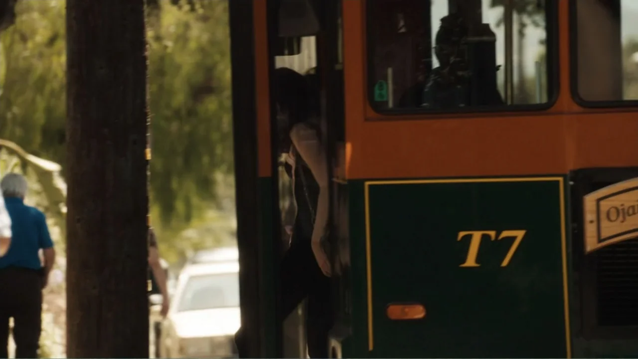 Easy A (2010) movie still: medium — A person exits a green and orange shuttle bus parked on a tree-lined street. The individual steps onto the curb…