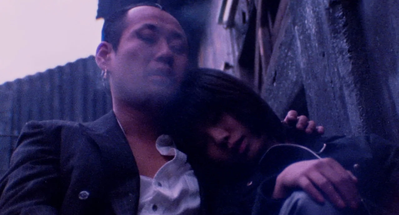 Burst City (1982) movie still: medium — An urban alleyway features dark metallic surfaces and textured concrete walls at night. A man and woman rest…