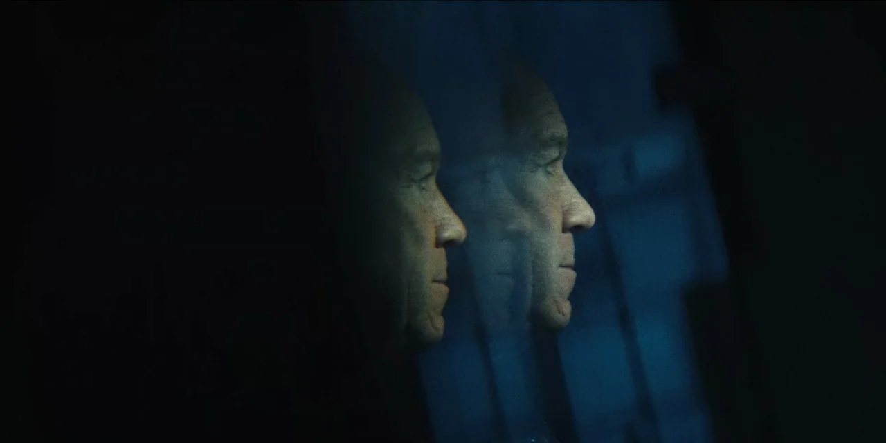 Spermworld (2024) movie still: closeup — A dark, interior setting features a reflection of a man in glass. The man looks out into the night through…