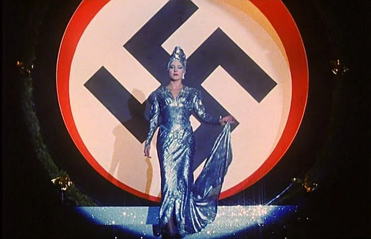 Lili Marleen (1981) movie still: fullbody — A woman in a blue gown stands on a stage before a swastika emblem. The performer pauses at the center of…