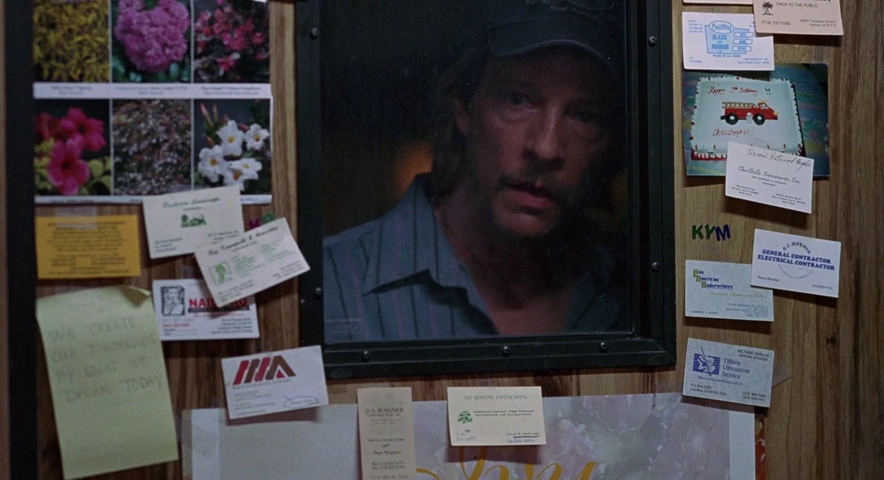 Adaptation (2002) movie still: medium — A wooden wall covered in various business cards, flyers, and a birthday drawing. A man looks through a small…
