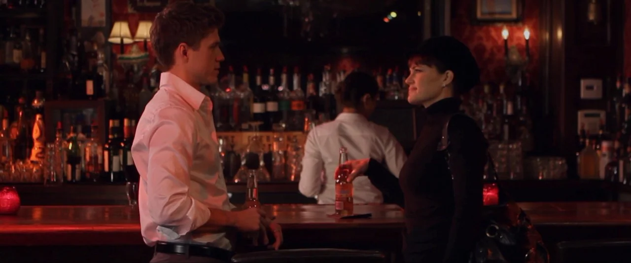 Girl Walks Into a Bar (2011) movie still: medium — A dimly lit bar features dark red walls with rows of illuminated spirit bottles. A man in a white…