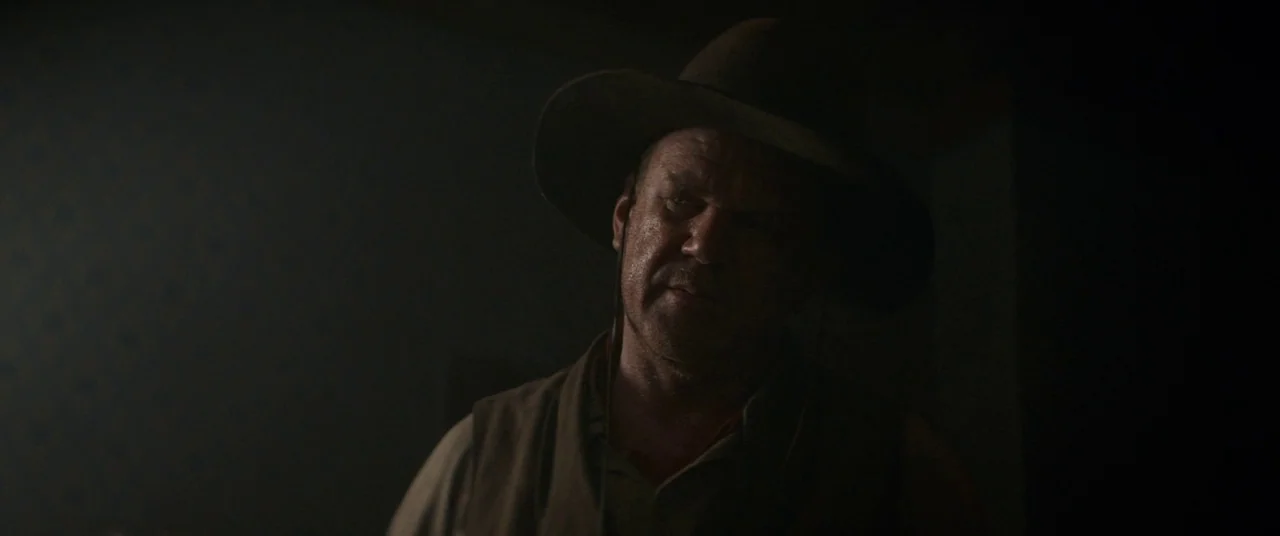 The Sisters Brothers (2018) movie still: medium — A dark, dimly lit room with minimal visible detail in the shadows. A man wears a wide-brimmed hat…