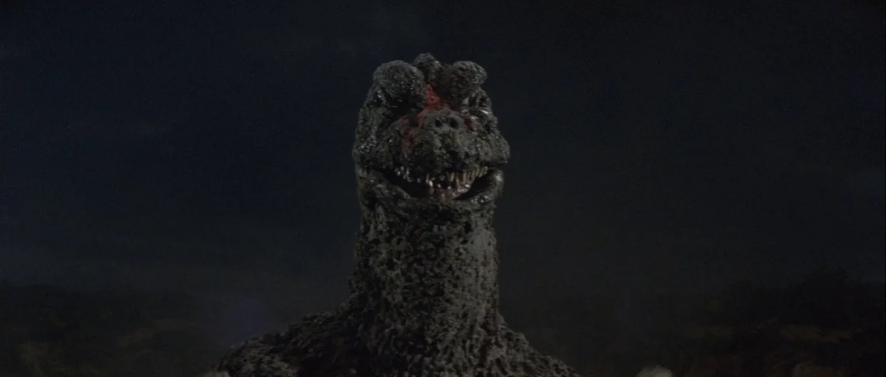 Godzilla Vs Gigan (1972) movie still: closeup — A dark night scene shows the head and neck of a large reptilian monster. The creature faces the…
