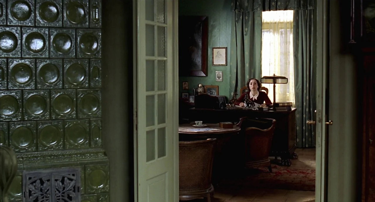 The Pianist (2002) movie still: wide — A study features olive green walls decorated with framed pictures and a tile stove. A woman sits behind a…