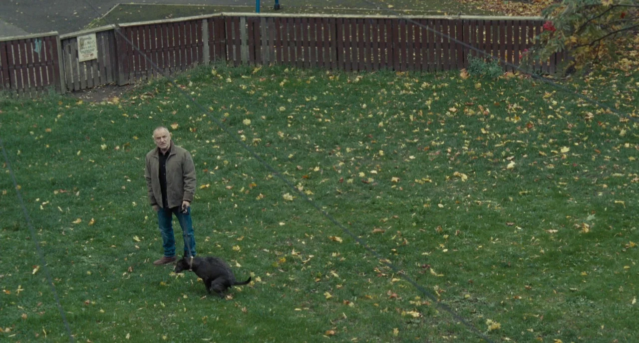 I, Daniel Blake (2016) movie still: wide — An enclosed public park with green grass littered with scattered autumn leaves. An older man stands…