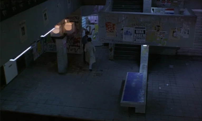 Spetters (1980) movie still: wide — A dimly lit concrete subway station interior features graffiti-covered walls and posters. Two figures walk away…