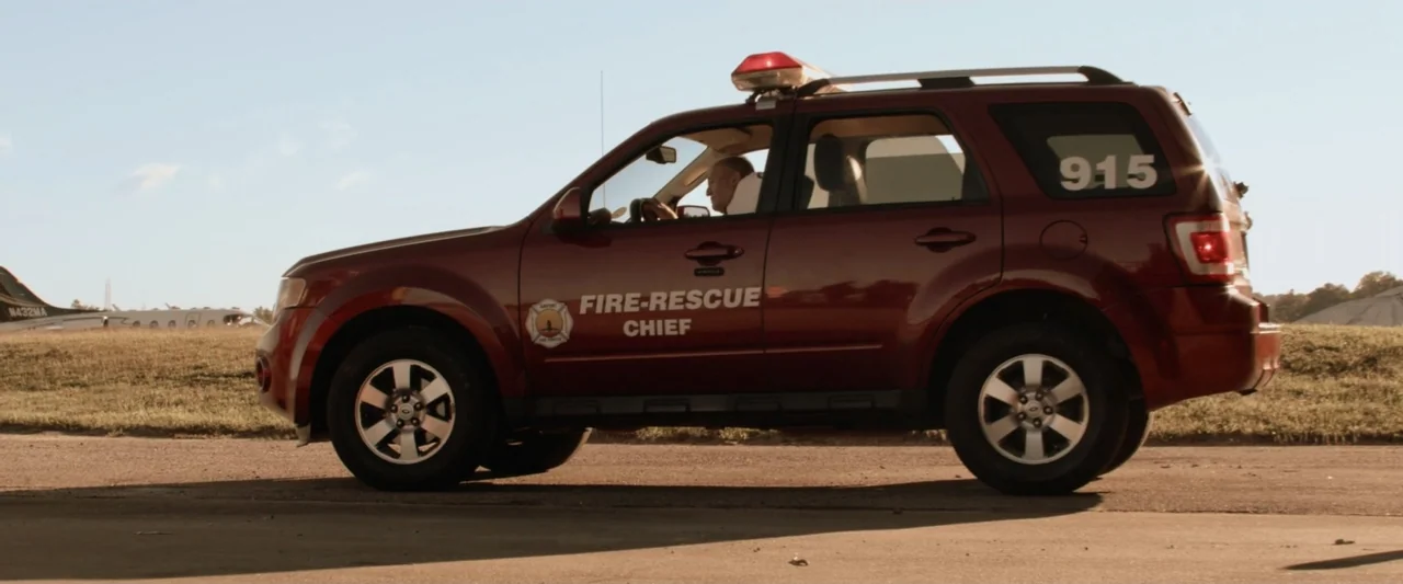 On a Wing and a Prayer (2023) movie still: medium — A deep red emergency vehicle drives across an open asphalt area near a runway. The driver…