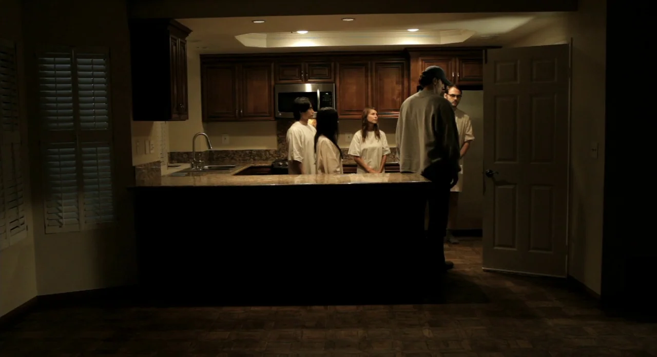Sound Of My Voice (2011) movie still: wide — A residential space contains dark wood cabinets above a granite countertop. Five individuals stand in a…