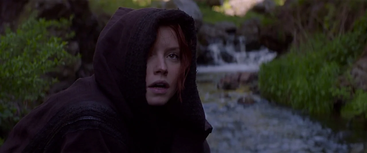 Ophelia (2018) movie still: closeup — A rocky stream flows through a lush green landscape with thick foliage. A young woman in a dark hooded cloak…