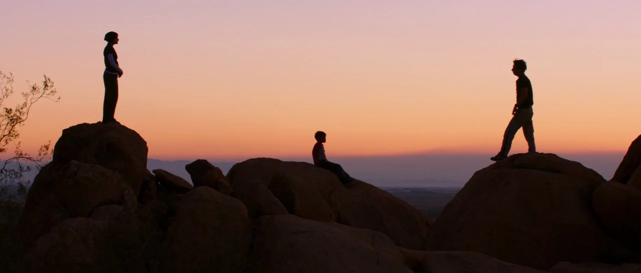 Wish I Was Here (2014) movie still: wide — Large boulders are situated on a desert hill during the twilight hours. Two adults and a child stand and…