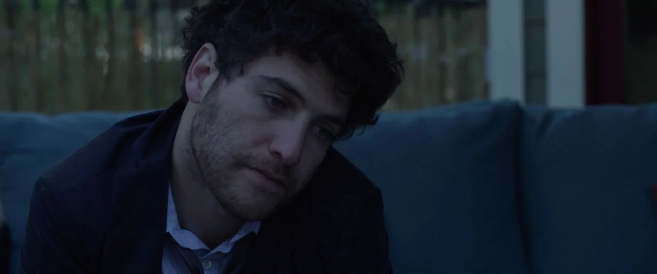 Night Owls (2015) movie still: closeup — An outdoor patio area at dusk features a plush sofa against a blurred fence. A man in a dark suit sits on…