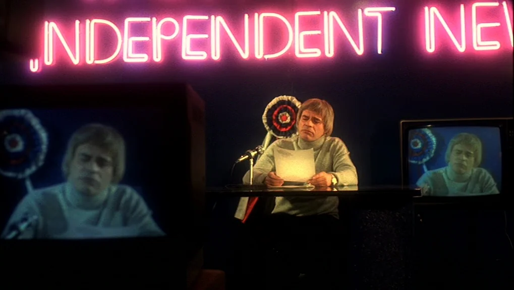 The War of the Worlds: Next Century (1981) movie still: medium — A television news studio features a bright pink neon sign mounted on a dark wall. A…