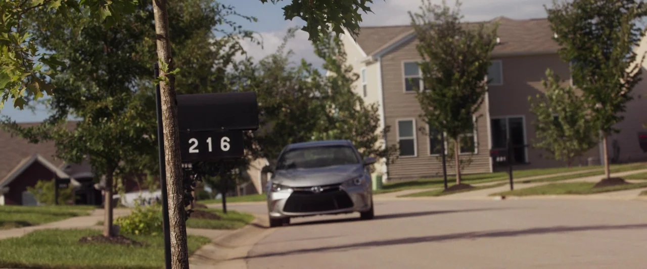 Mom and Dad (2017) movie still: wide — A residential suburban street features houses with vinyl siding and manicured green lawns. A silver sedan is…