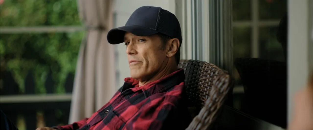 Someone Like You (2024) movie still: medium — A man sits on a patio in a wicker chair near a window. The man wears a plaid shirt and cap while…