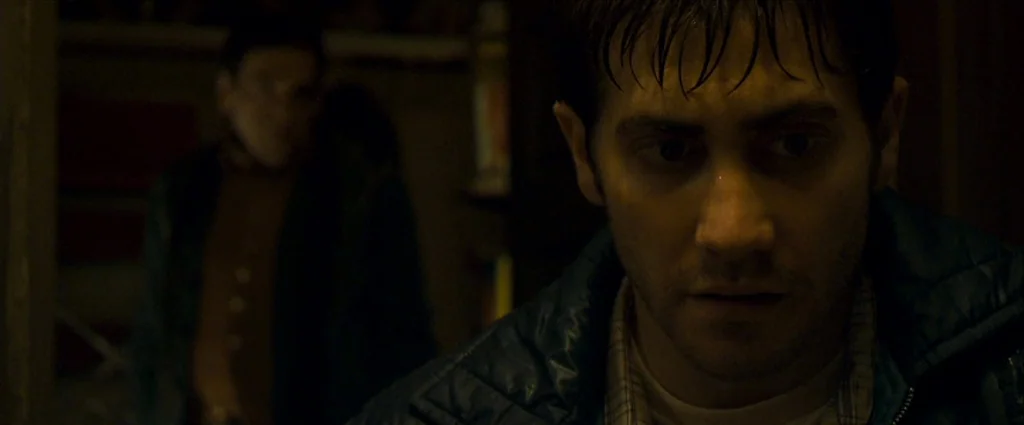 Zodiac (2007) movie still: closeup — A dimly lit interior space with a secondary figure blurred in the deep background. The main subject is in the…