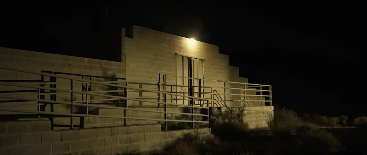 American Sicario (2021) movie still: wide — A concrete block building with a metal railing sits in a dark desert landscape. A single light…