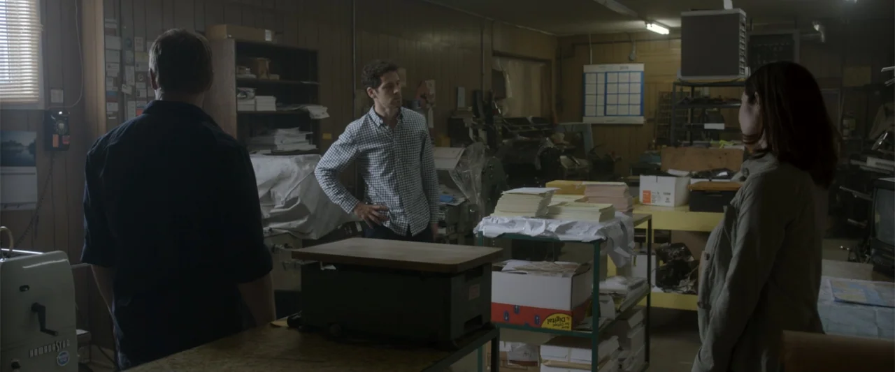 Radius (2017) movie still: medium — A cluttered print shop office with wooden paneling, heavy machinery, and paper supplies. Three adults stand near…