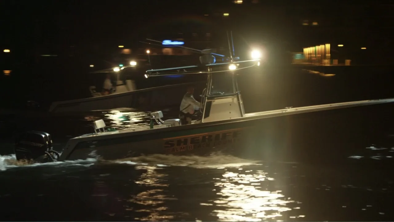 Parker (2013) movie still: medium — A law enforcement boat moves across dark water at night under bright overhead lights. An officer in a uniform…