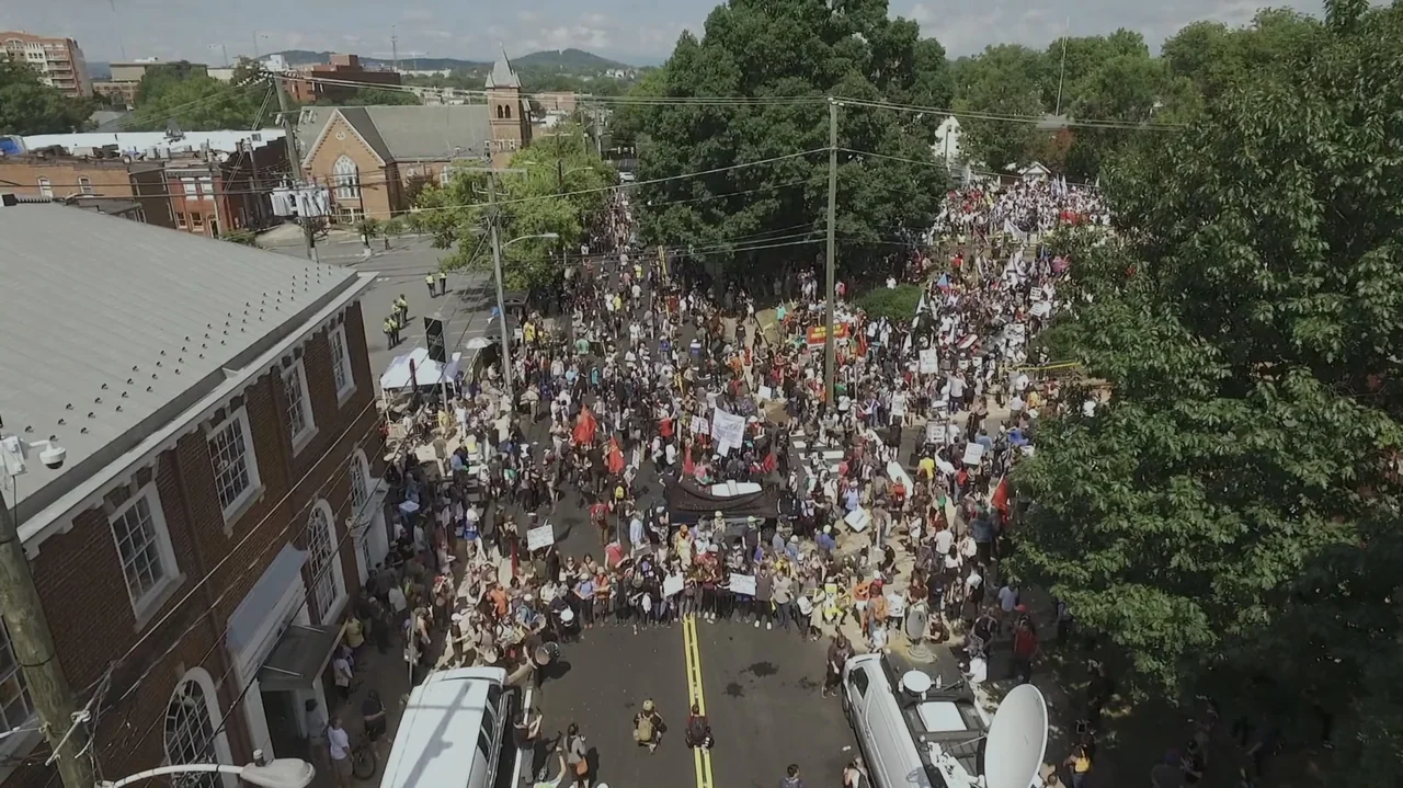 The Soul of America (2020) movie still: establishing — A dense crowd of people occupies a park area near a large brick building. Participants hold…