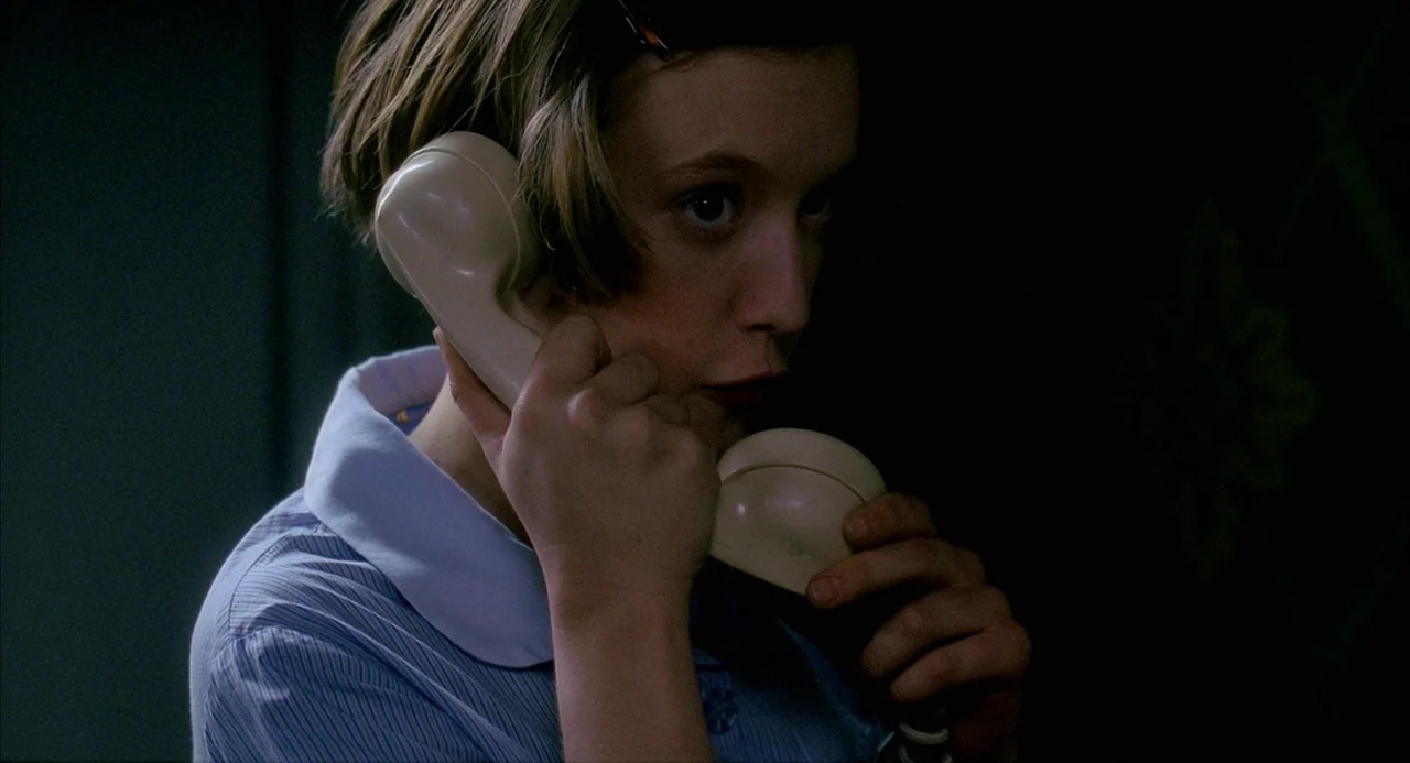 8 Women (2002) movie still: closeup — A dark space provides a minimal backdrop for a woman at a telephone. A woman with a short bob hairstyle holds…