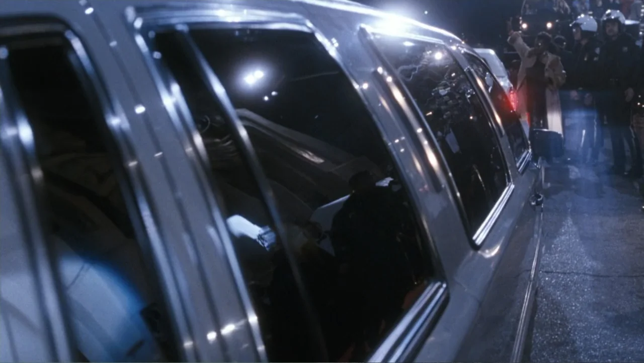 The Bodyguard (1992) movie still: medium — A silver luxury vehicle sits parked on an asphalt city street at night. Police officers in uniform stand…