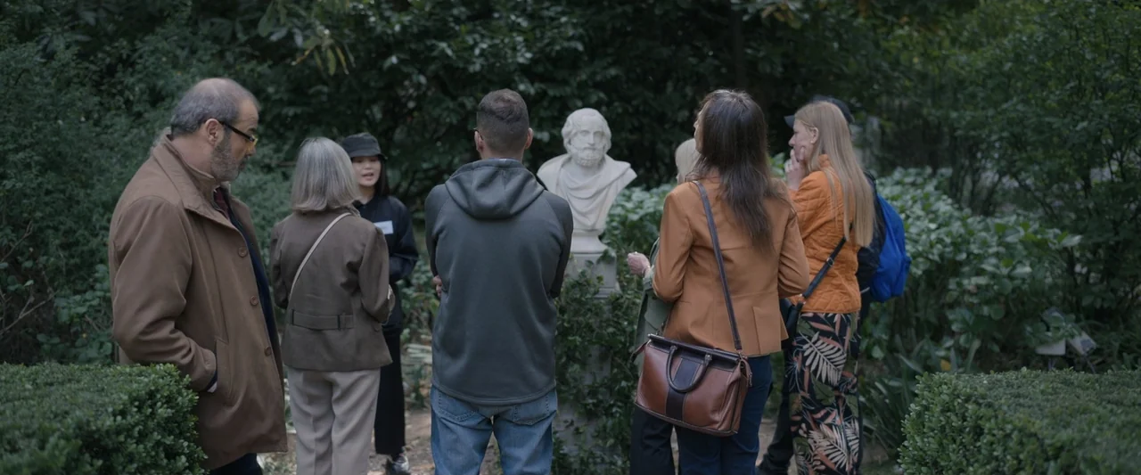 Doublespeak (2021) movie still: fullbody — A group of people gathers in a lush, green outdoor space around a sculpture. They stand together while…