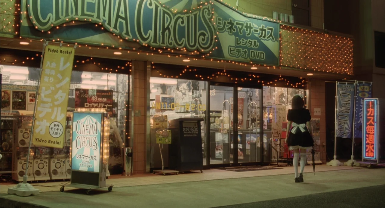 Air Doll (2009) movie still: fullbody — A Japanese video rental shop front at night with lit signs and banners. A person in a maid costume walks…