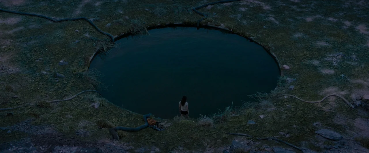 Settlers (2021) movie still: wide — A woman stands alone at the edge of a large, man-made circular concrete pool. Several black plastic pipes snake…