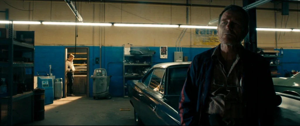 Drive (2011) movie still: medium — An auto repair shop features blue painted walls and metal storage shelves with clutter. A man in a dark jacket…