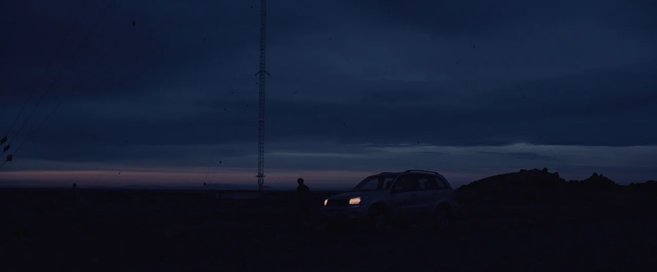 Rift (2017) movie still: wide — A dark, expansive field at dusk features a tall telecommunications tower in the background. A single person stands…