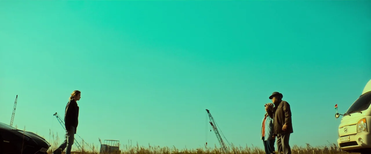 Ballerina (2023) movie still: wide — A flat, grassy field contains two parked vehicles and several construction cranes in distance. A woman walks…