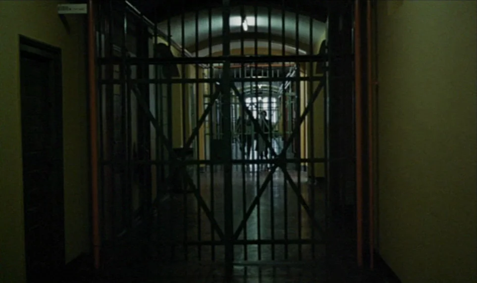 Stroszek (1977) movie still: establishing — A long, dimly lit indoor corridor behind a heavy black iron security gate. The yellow-walled hallway…