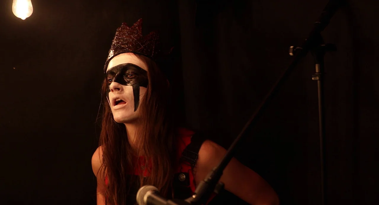 Hellbender (2021) movie still: medium — A dark performance area with a black backdrop and a single hanging lightbulb. A person with black face paint…
